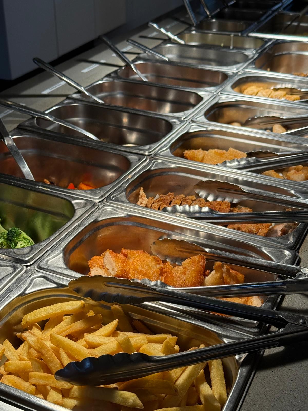 Buffetzeile mit Edelstahlbehältern: Pommes frites, frittiertes Hähnchen, Brokkoli und weitere warme Gerichte, jeweils mit Zange.