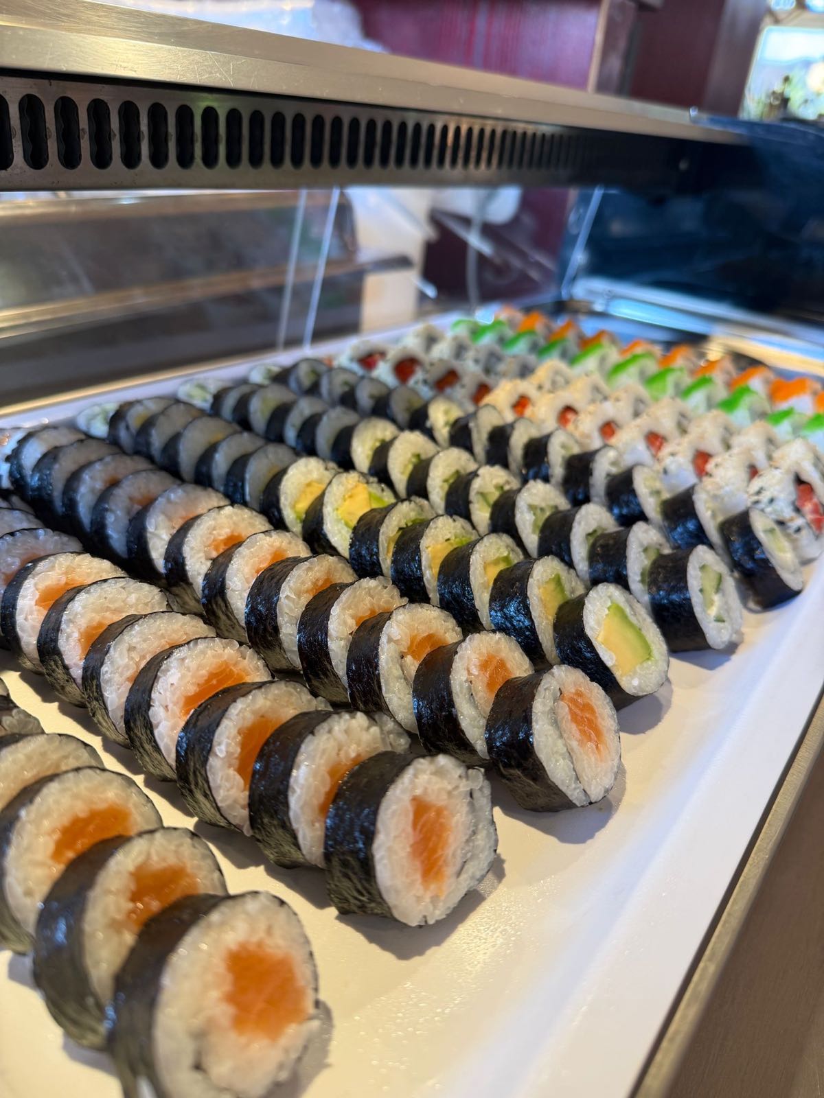 Sushi-Theke: Reihen frisch gerollter Maki mit Lachs, Avocado, Gurke und Ei in der gekühlten Vitrine.